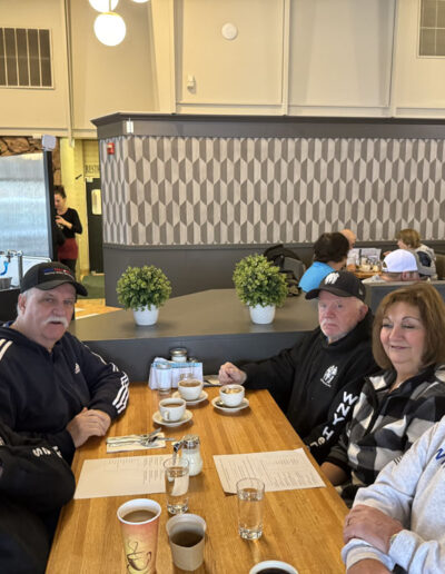Five smiling veterans and supporters seated around a wooden diner table with coffee cups and menus, several wearing WNY Heroes and military-related clothing, inside a warmly lit restaurant.