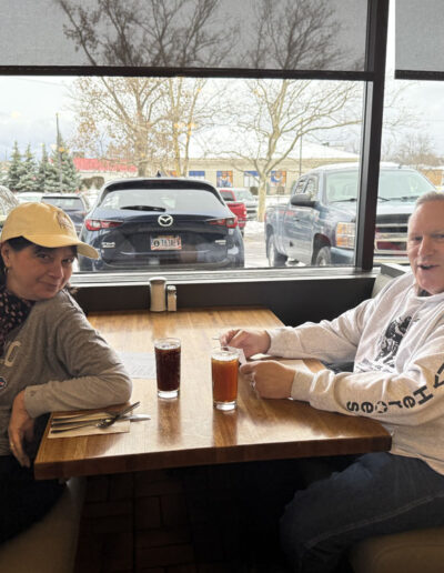 Two people sitting across from each other in a diner booth, woman wearing a Buffalo Bills sweatshirt and yellow cap, man in a gray hoodie holding a glass, winter parking lot outside the window.