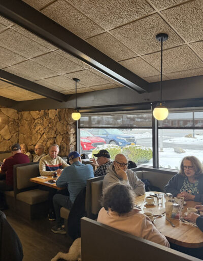 Wide view inside a cozy diner filled with veterans and supporters having breakfast together on a winter morning, snowy parking lot visible through large windows.