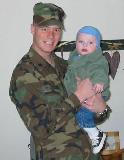 Heath in Army camouflage uniform proudly holding his young child, both smiling at home before deployment.