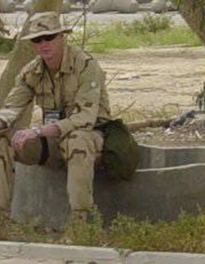 Heath in desert camouflage sitting outdoors on a concrete barrier under a tree, relaxed moment during deployment.