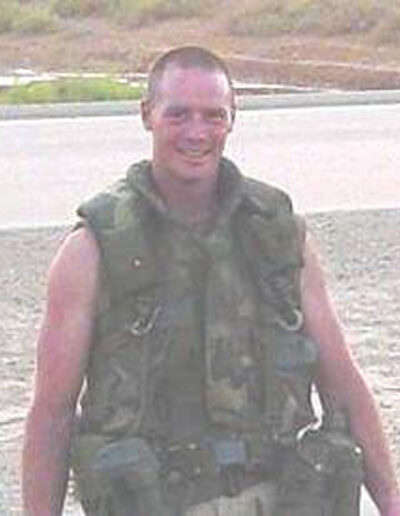 Heath in full combat gear and body armor standing outdoors during deployment, smiling slightly at the camera.