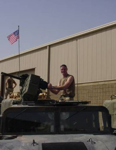 Heath in desert tan uniform standing atop a Humvee turret with a mounted machine gun, American flag flying behind him in Iraq.