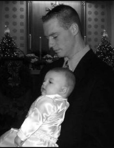 Heath gently cradling his infant son at a Christmas baptism or christening, surrounded by softly lit Christmas trees, black-and-white photo.