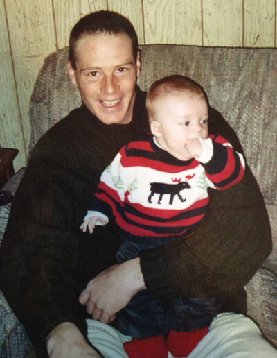 Heath McMillin smiling warmly while holding his young son on his lap, both dressed in cozy winter clothes, seated on a couch with wood-paneled walls.