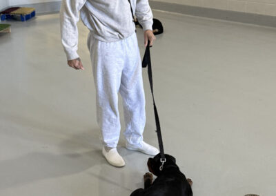 Man in gray prison uniform and black cap stands in a training room while a black dog lies calmly in a perfect “down” position in front of him.