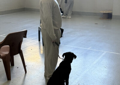 Man in gray prison uniform and orange beanie stands in a training room holding the leash of a black dog sitting perfectly at his side.