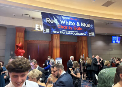 Entrance area of the WNY Heroes Red, White & Blue Charity Gala with a large hanging banner, red-white-blue balloons, and formally dressed guests arriving or socializing.
