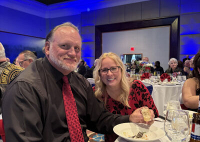 Smiling couple seated at a round banquet table during the WNY Heroes gala dinner, man with beard wearing a red tie, woman in a red and black dress holding food.