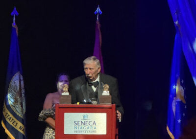 Older man with beard speaking at the Seneca Niagara podium while a woman in a gold and black gown stands nearby at the WNY Heroes Red, White & Blue Gala.