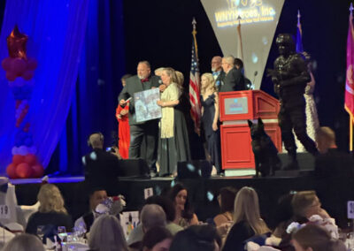 On-stage award presentation at the WNYHeroes gala: a man hands a large framed item to a woman while others watch near a red podium and military working dog statue.