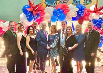 Nine people in formal attire pose in front of a red-white-blue balloon arch and large illuminated “2025” letters at a gala.