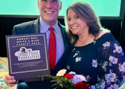 A man and woman smile on stage holding an award plaque for “The Jim Fiore Foundation” and a bouquet of red and white roses in front of the WNYHeroes backdrop.