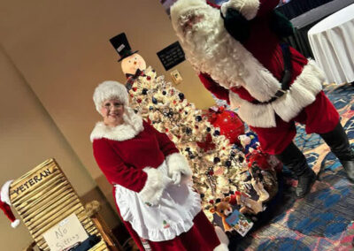 Mr. and Mrs. Claus pose cheerfully in front of a patriotically decorated Christmas tree and a “VETERAN No Sitter” chair at a festive veterans event.