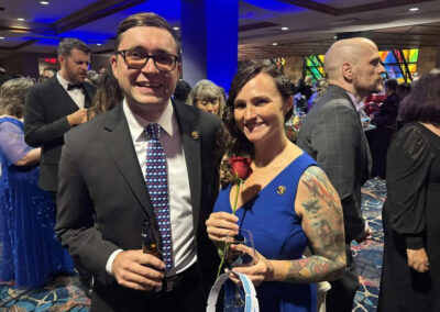 A man in a suit and a woman in a blue dress with visible tattoos smile while holding drinks and a red rose at a crowded formal gala event.