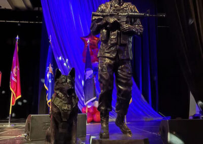 Bronze statue of a soldier with rifle and a military working dog displayed on stage under blue lighting, flanked by American and service flags.