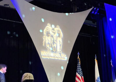 A man on stage holding papers stands beside a battlefield cross memorial (helmet on rifle) with the WNYHeroes logo projected behind and American flags on either side.