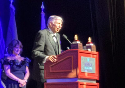 An older man in a suit speaks at a red Seneca Niagara podium while a woman in a black dress stands beside him during an awards segment.