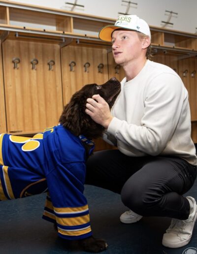 A Sabres player wearing a white “OILERS 47” cap and cream sweatshirt kneels down in the locker room to gently pet Mac, a brown Newfoundland puppy wearing a classic blue-and-gold Sabres jersey, who is gazing up adoringly.