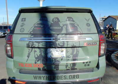 Rear view of a green Chevrolet SUV wrapped with “WNYHeroes.org” and “Pawsitive for Heroes” veteran and service-dog graphics, parked outdoors.