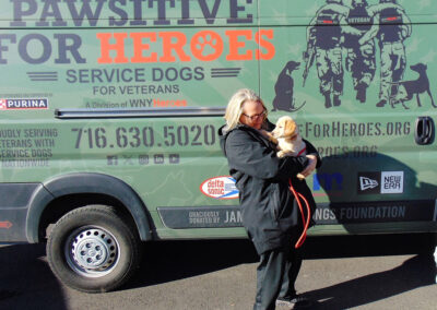 A woman in a black jacket stands smiling next to the “Pawsitive for Heroes” service-dogs-for-veterans van, cradling a young yellow Labrador puppy.