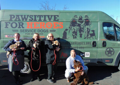 Four people stand outdoors in front of a green “Pawsitive for Heroes” van, each holding a puppy (three yellow Labs and one brown Newfoundland).