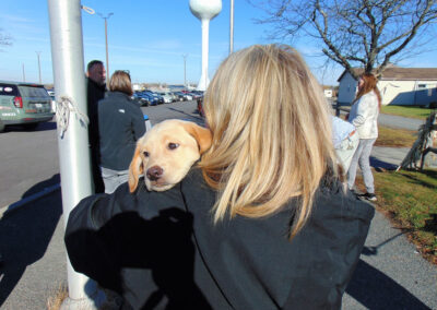 A woman with long blonde hair holds a young yellow Labrador puppy over her shoulder outdoors on a sunny day, with parked cars and a water tower in the background.