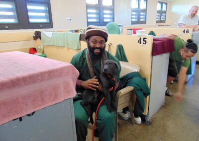 An incarcerated man in green uniform and knit hat sits smiling in a dormitory-style housing unit, gently holding a small black puppy in his lap.
