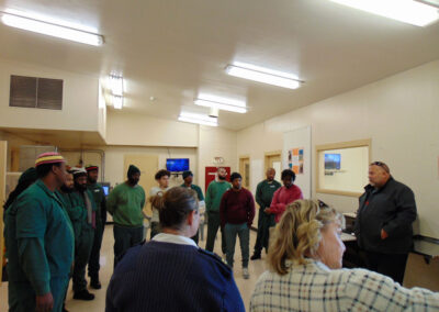 A group of incarcerated men in green uniforms and a few staff members gather in a circle inside a jail common room, listening to two women speaking.