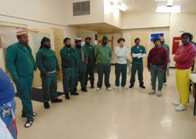 Nine incarcerated men in green prison uniforms stand in a loose line inside a brightly lit common area of a correctional facility, facing the camera.