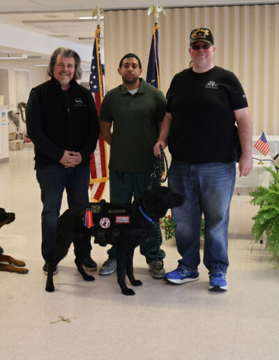 Three men indoors: one in a green prison uniform stands between two staff members; a black service dog in training wearing a vest stands calmly in front of them, with American and New York state flags in the background.