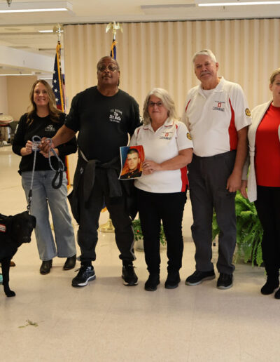 Three men indoors: one in a green prison uniform stands between two staff members; a black service dog in training wearing a vest stands calmly in front of them, with American and New York state flags in the background.