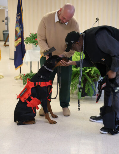Eight people (a mix of incarcerated men in green uniforms and program staff/volunteers) stand smiling indoors with two service dogs in training, one black and one tan-and-white, in front of American and state flags.