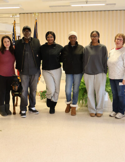 Eight people (a mix of incarcerated men in green uniforms and program staff/volunteers) stand smiling indoors with two service dogs in training, one black and one tan-and-white, in front of American and state flags.