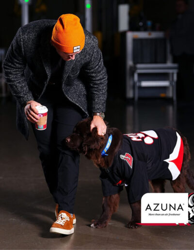 A Sabres player in an orange beanie and dark coat holds a coffee cup in one hand while petting Mac, a brown Newfoundland puppy in a black Sabres jersey with a big red paw print, standing on the rink floor. Azuna logo in corner.