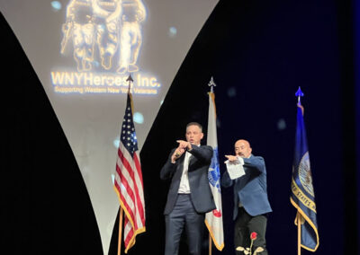 Two men in suits on stage at the WNYHeroes Red, White & Blue Gala, one speaking into a microphone and gesturing while the backdrop displays the WNYHeroes logo and American flags.