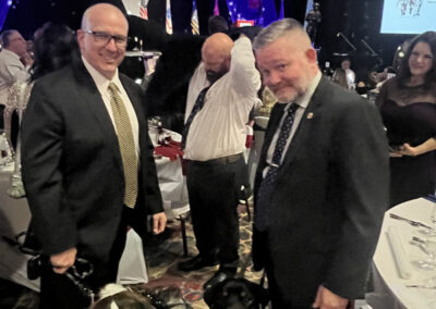 Three men in suits pose with two service dogs (a brown-and-white spaniel mix and a black-and-tan Rottweiler) at a formal gala dinner with patriotic stage lighting.