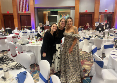 Three women in formal attire (black velvet, patterned skirt, and metallic gold gown) smile and pose together in a banquet hall decorated with red, white, and blue chair sashes and an American flag on the wall.