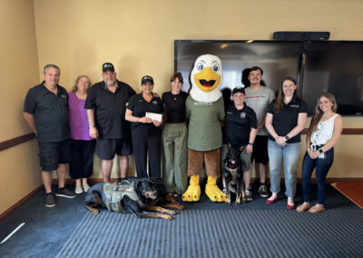 A group of nine smiling people from WNYHeroes and community partners pose together in a conference room with an eagle mascot in a green WNYHeroes shirt. Two service dogs in tactical vests (one Rottweiler lying, one sitting) are in the front row. Someone is holding a ceremonial check.